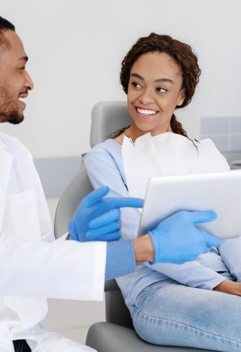 Cheery african guy dentist showing woman patient his clinic website on digital tablet